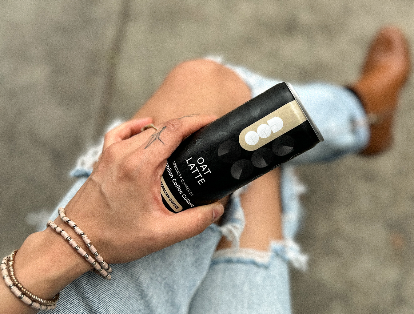 Person holding a black and gold 'Oat Latte' container with denim jeans and a blurred background